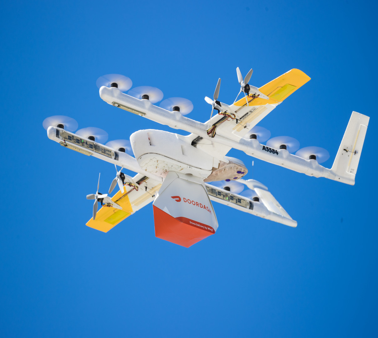A Wing delivery aircraft carrying a DoorDash package in flight against a deep blue sky.