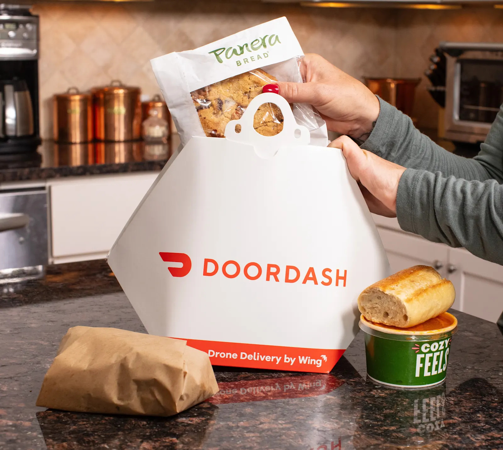 Hands lift a Panera Bread cookie out of a DoorDash delivery box marked Drone Delivery by Wing.