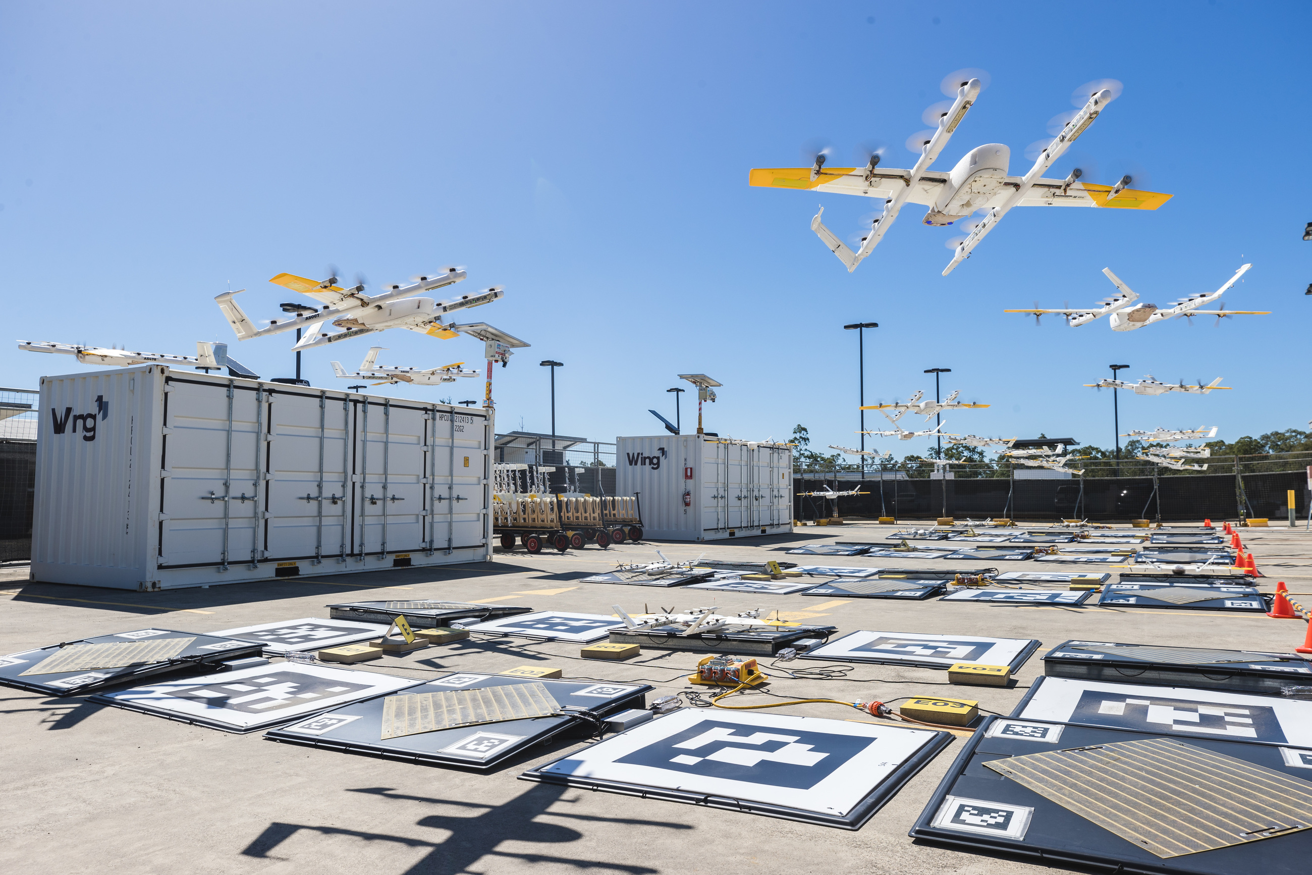 A fenced operating site: rows of marked charging pads on asphalt, shipping containers, and delivery aircraft in the air above.