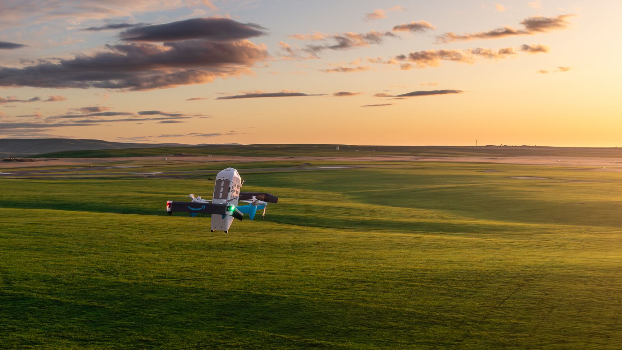 An Amazon Prime Air aircraft in flight low over open green fields at sunset.