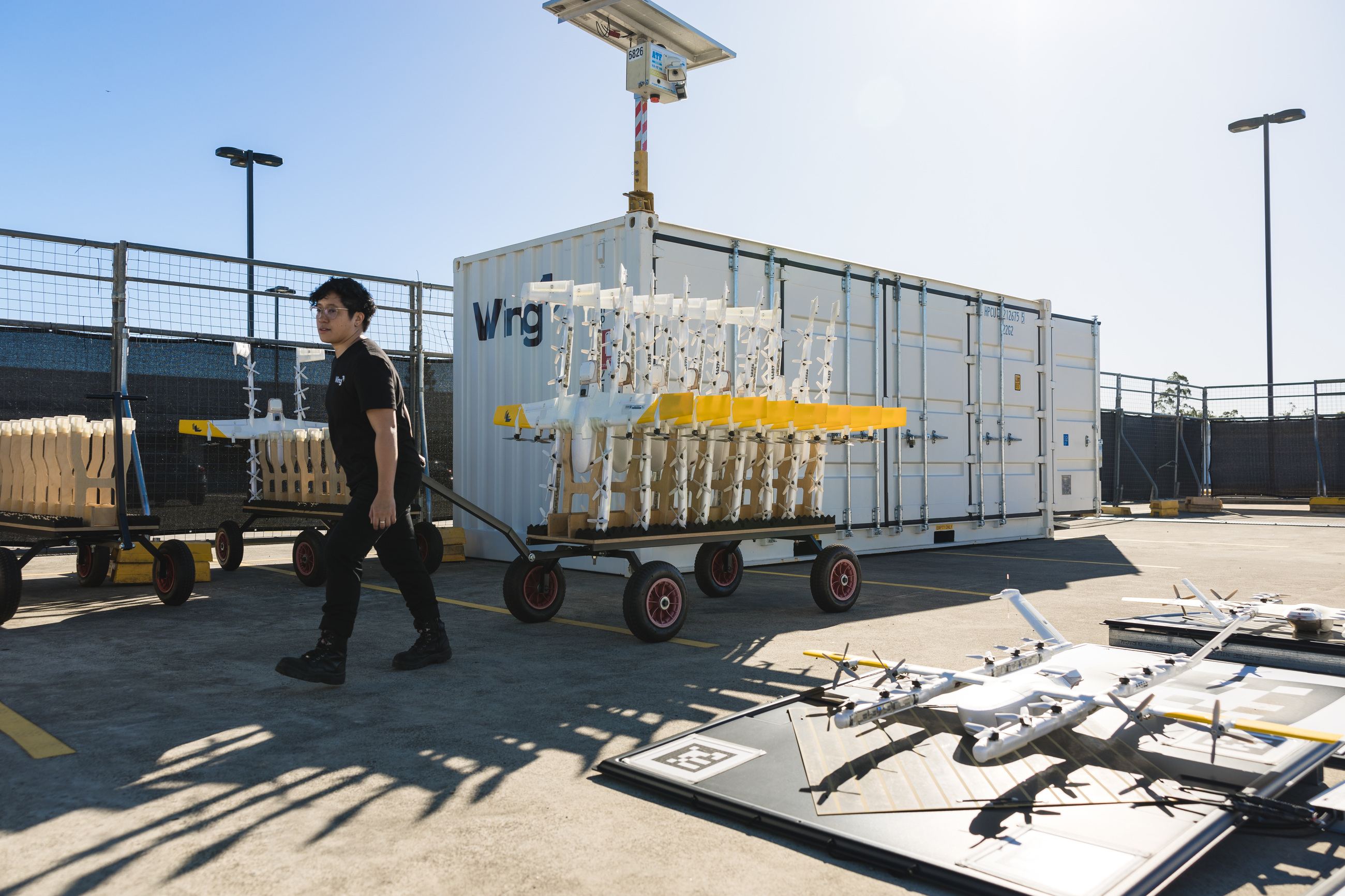 A technician wheels a rack of aircraft past a shipping container inside a fenced operating site.