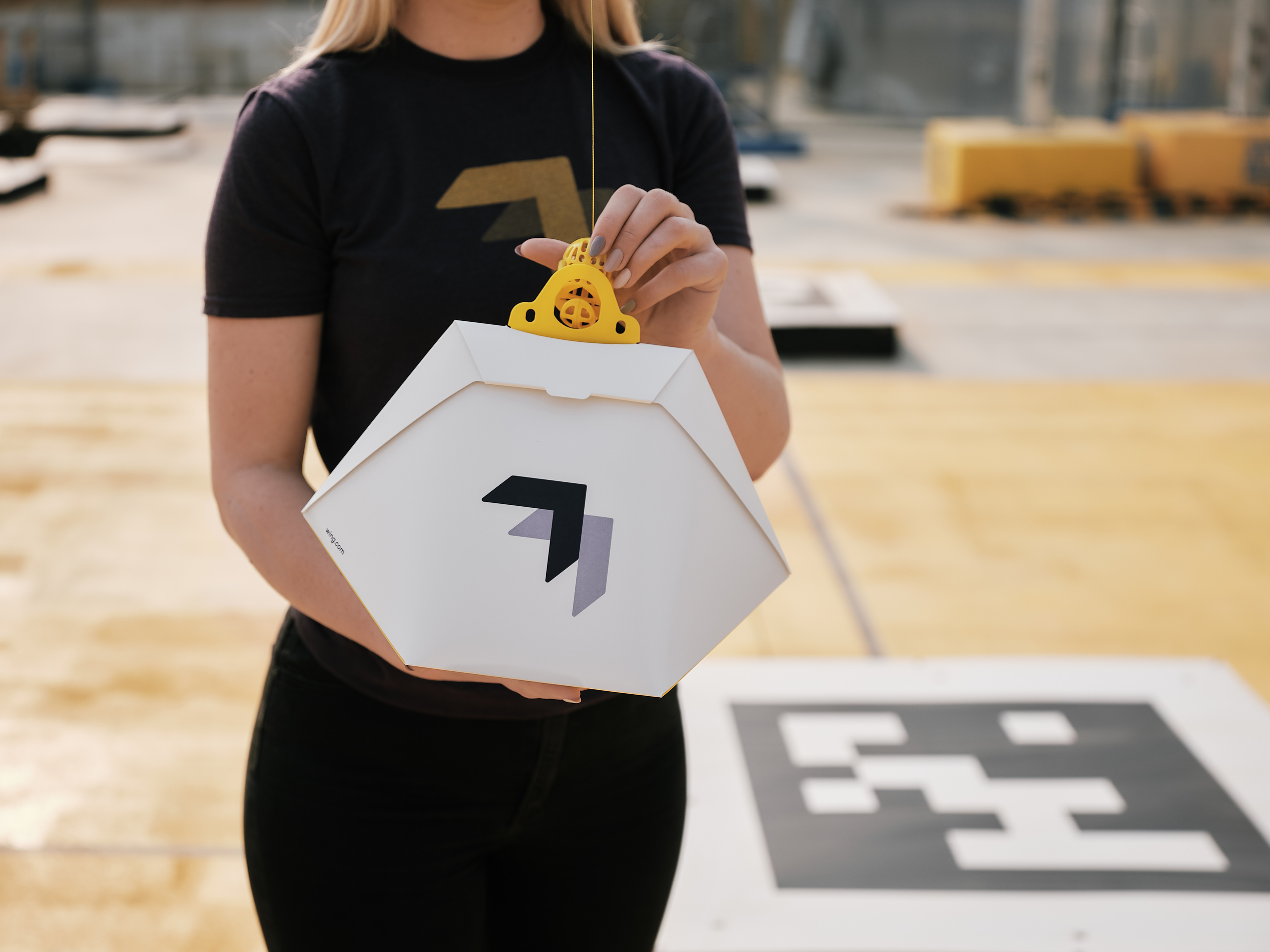 A Wing employee clips a delivery box to the aircraft tether beside a marked charging pad.