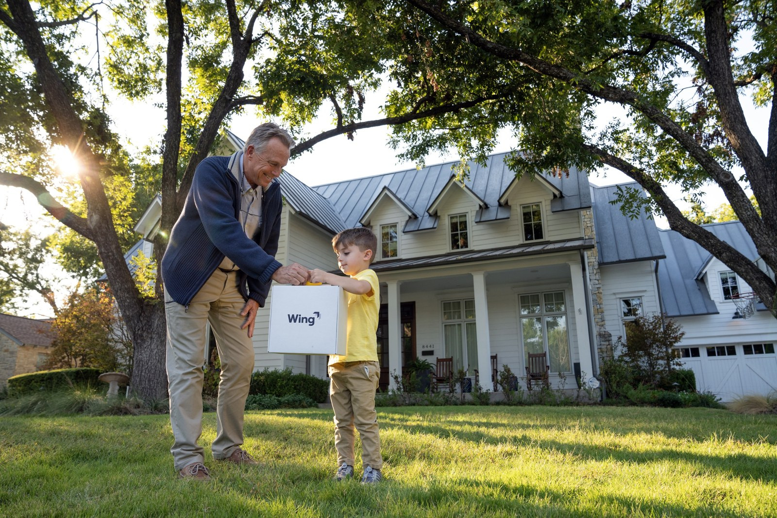 An adult and a young child open a delivery box together on a front lawn in late afternoon light.
